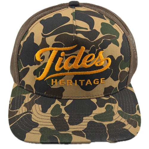 A trucker-style cap featuring a green and brown camouflage pattern on the front panel and brim, with a dark brown mesh back. The front is embroidered with 'Tides HERITAGE' in yellow-gold thread.  THE BRAND IS TIDES HERITAGE AND FEATURES A DUCK CAMO.  